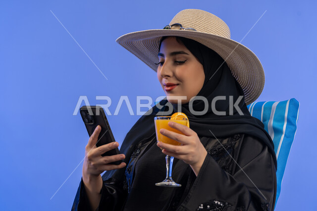 الاستمتاع بالمشروبات الباردة وقضاء أيام العطلة في الرحلات السياحية , بورترية مقرب لامراة عربيه خليجيه امارتيه محجبة ترتدي العباءه السوداء و قبعه من القش, امراة سعودية مبتسمة تقوم بالنظر فى الهاتف المحمول بجانب حافظة التبريد , خلفية زرقاء