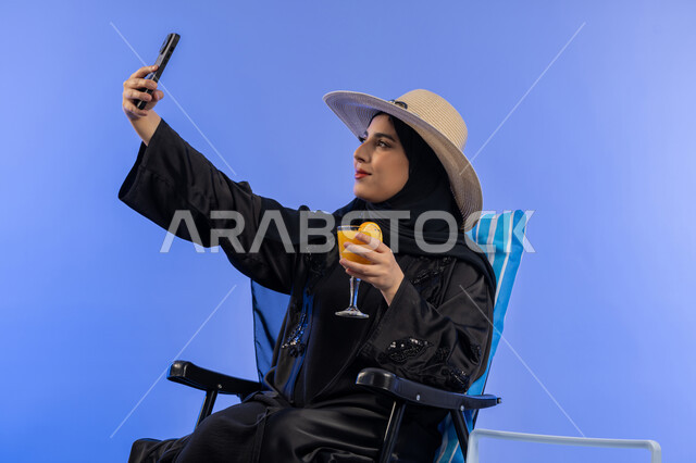 الاستماع بالمشروبات الصيفية المنعشة, بورتريه لشابة عربية خليجية اماراتية مبتسمة ترتدي العباءة السوداء و قبعة القش تقوم بالتقاط صور سيلفي تذكارية عبر الهاتف المحمول, السفر والاستمتاع بالأجواء الصيفيه، خلفية زرقاء