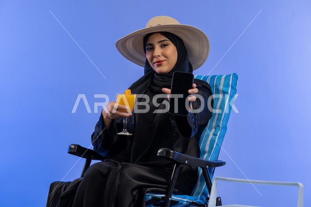 القيام بالجولات الترفيهية وقت الإجازة , بورترية لامراة عربيه خليجيه اماراتية محجبة ترتدي العباءه السوداء و قبعه من القش تجلس على كرسي مريح, سيدة سعوديه تقوم بعرض شاشه جوال سوداء و في يديها كوب عصير منعش, خلفية زرقاء