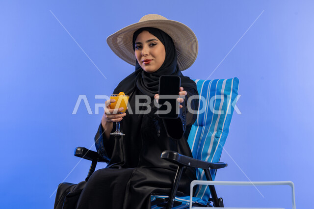 قضاء وقت ممتع فى الإجازة, بورترية لامراة عربيه خليجيه اماراتية محجبة ترتدي العباءه السوداء و قبعه من القش تجلس على كرسي مريح, سيدة سعوديه تقوم بعرض شاشه جوال سوداء و في يديها كوب عصير منعش, خلفية زرقاء