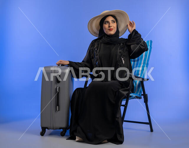 مفهوم الرحلات السياحية, بورترية لامراة عربيه خليجيه اماراتية محجبة ترتدي العباءه السوداء تنظر الى شيء ما, سيدة سعوديه مبتسمه تمسك قبعه القش بيديها بجانب حقيبه السفر , قضاء أوقات ممتعة, خلفية زرقاء
