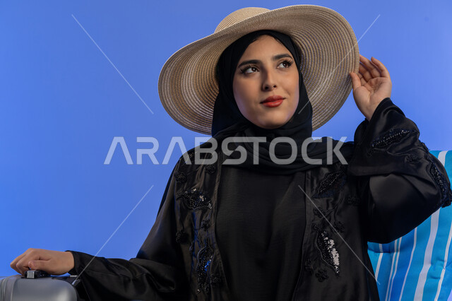 الشعور بالاستجمام والفرح, بورترية مقرب لامراة عربيه خليجيه اماراتية محجبة ترتدي العباءه السوداء تنظر الى شيء ما, سيدة سعوديه مبتسمه تمسك قبعه القش بيديها بجانب حقيبه السفر , قضاء أوقات ممتعة, خلفية زرقاء