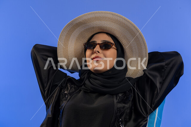 قضاء اوقات ممتعه فى العطله الصيفية , بورترية مقرب لامراة عربيه خليجيه امارتيه محجبه ترتدي العباءه السوداء و قبعه من القش, سيده سعودية تجلس مسترخية على كرسي بجانب حقيبة السفر,الاستمتاع بالعطلة الصيفية لاستعادة الحيوية والنشاط، خلفية زرقاء