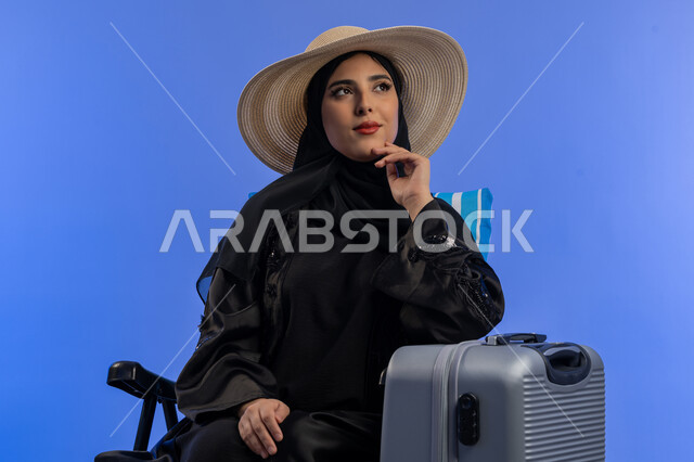 الشعور بالسعادة والفرح ، الاستمتاع بالعطلة الصيفية وقضاء وقت ممتع,بورتريه لامراة عربية خليجية اماراتية مبتسمة ترتدي الزي العباءة السوداء و قبعه القش بجانبها شنطة سفر  , سيدة سعوديه تنظر الى الاعلي و تفكر فى شيء ما, خلفية زرقاء