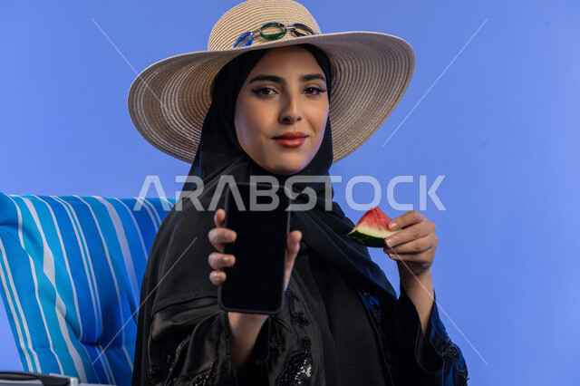 استخدام التكنولوجيا و التقنيات الحديثة, تناول الفواكه الصيفية المنعشة, بورتريه لامراة عربية خليجية اماراتية مبتسمة ترتدي العباءة السوداء و قبعه القش تستخدم الهاتف المحمول, إمرأة سعودية تقوم بعرض شاشه فارغه, الاستمتاع بالفاكهه الصيفية, خلفية زرقاء