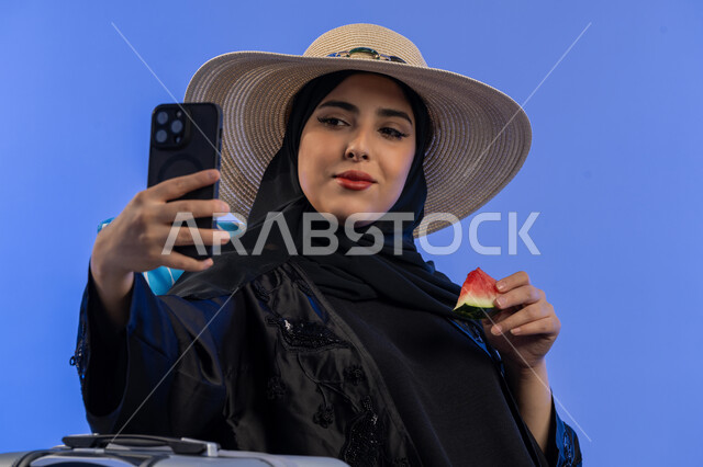 قضاء أيام ممتعة فى العطلة, تناول الفواكه الصيفية المنعشة, بورتريه مقرب لشابة عربية خليجية اماراتية مبتسمة ترتدي العباءة السوداء و قبعه القش تستخدم الهاتف المحمول, إمرأة سعودية تقوم بالتقاط صور سيلفي تذكارية عبر الجوال, خلفية زرقاء