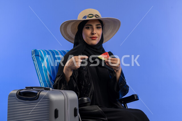 قضاء اوقات ممتعة فى الاجازة و تناول الفواكه الصيفية, بورترية لامراه عربية خليجيه اماراتية محجبة ترتدي العباءه السوداء و قبعه من القش تاكل قطعة من البطيخ, سيده سعوديه مبتسمه تجلس على كرسي بجانب حقيبه السفر , خلفية زرقاء