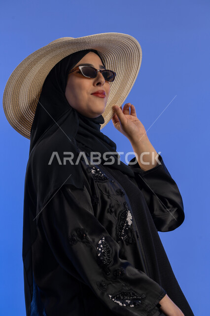 رحله سياحيه فى إجازه صيفيه و قضاء وقت ممتع، بورتريه لإمرأه عربية خليجيه امارتية محجبة ترتدي نظارة شمسية تقف بإيماءات وجه تدل على السعاده و الثقة, سيده سعوديه مبتسمة تضع يديها على قبعه القش و تنظر الى شيء ما، خلفيه زرقاء
