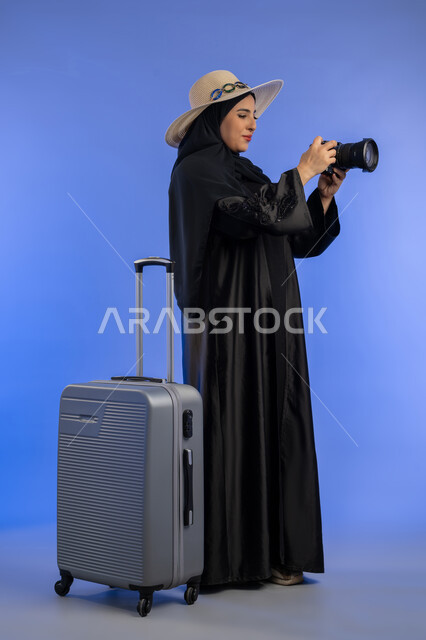 ممارسة هواية التصوير، بورتريه لإمراة عربية خليجية اماراتيه ترتدي الحجاب والعباءة تمسك بيدها كاميرا رقمية تقوم بالتقاط الصورالتذكارية بجانب شنطة سفر، سيده سعوديه تستعد للسفر و قضاء العطله الصيفية, صورة لكامل الجسم, خلفية زرقاء