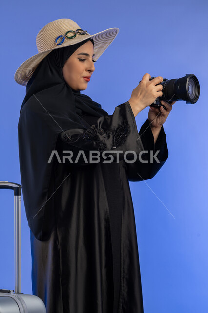 ممارسة هواية التصوير بتعابير السرور، بورتريه لإمراة عربية خليجية اماراتيه ترتدي الحجاب والعباءة تمسك بيدها كاميرا رقمية تقوم بالتقاط الصور التذكارية بجانب شنطة سفر، سيده سعوديه تستعد للسفر و قضاء العطله الصيفية, خلفية زرقاء