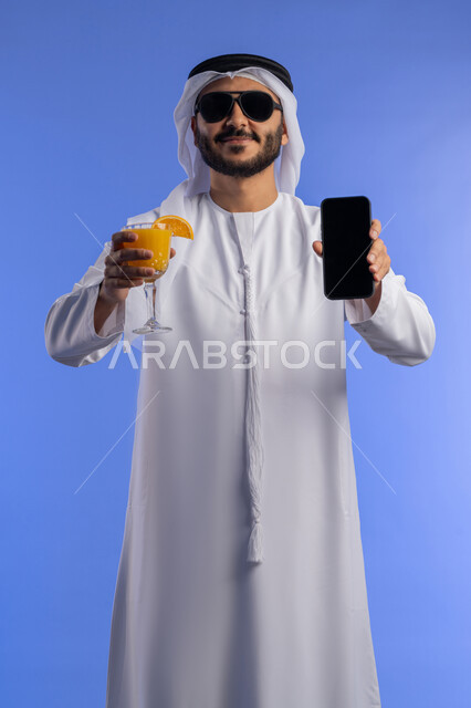 الاستمتاع بالمشروبات الصيفية المنعشة , الاستعداد لرحلة ترفيهية ممتعة, عرض شاشة سوداء فارغة على الموبايل, قضاء أوقات العطلة الصيفية فى السياحة والسفر, بورتريه لشاب عربي اماراتي يرتدي الزي التقليدي و يقوم بعرض الهاتف المحمول , خلفية زرقاء