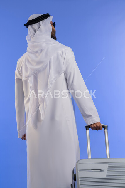 القيام بالجولات الترفيهية فى وقت الإجازة, تعابير الفرح والسرور, مفهوم السياحة فى إجازة الصيف، القيام بالجولات الترفيهية، بورتريه من الخلف لشاب عربي اماراتي يرتدي الزي التقليدي ويمسك بيده حقيبة الامتعة, خلفية زرقاء