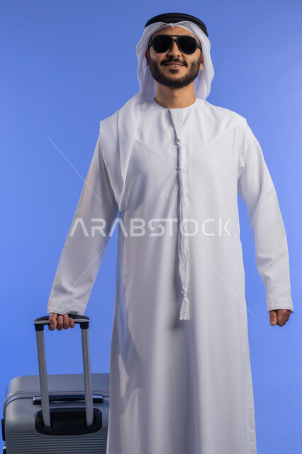 ايماءات السرور والمتعة والحماس, التسلية والترفيه فى الخارج، بورتريه لشاب عربي خليجي اماراتي مبتسم يرتدي الزي التقليدي والنظارة الشمسية يمسك بيده حقيبة السفر, الاستعداد لقضاء رحلة صيفية ممتعة، خلفية زرقاء
