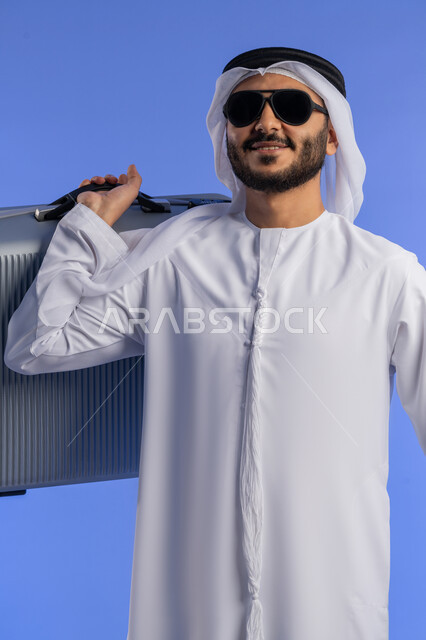 مفهوم السياحة والقيام بالجولات الترفيهية فى وقت الإجازة, تعابير الفرح والاستعداد لقضاء رحلة صيفية ممتعة، القيام بالجولات الترفيهية، بورتريه لرجل عربي خليجي اماراتي يرتدي الثوب التقليدي يحمل بيده حقيبة الأمتعة، خلفية زرقاء