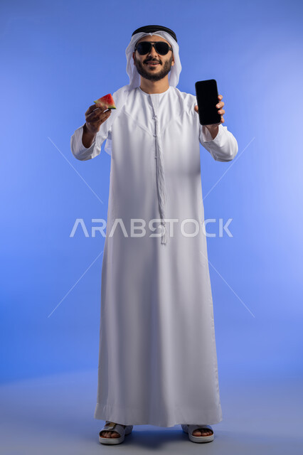 النظر الى الكاميرا بإيماءات السرور والسعادة، عرض هاتف محمول بشاشة سوداء فارغة، الاستمتاع بالفاكهه الصيفية الممتعة, بورتريه لرجل عربي خليجي اماراتي يرتدي الثوب التقليدي يمسك بيده قطعه من البطيخ و جهاز الجوال, خلفية لكامل الجسم, خلفية زرقاء
