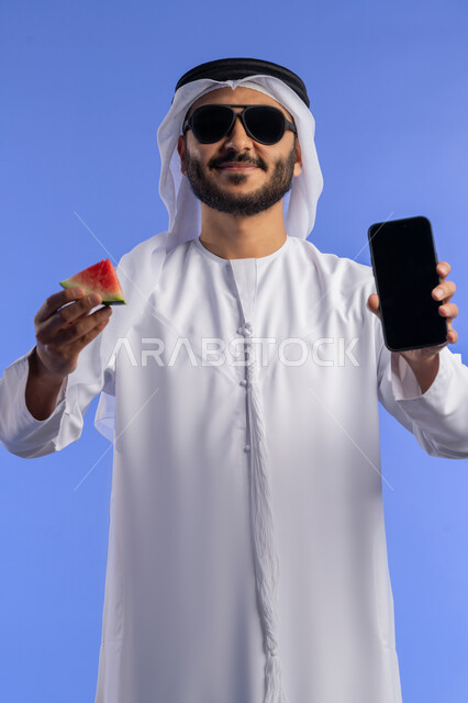 السياحة فى إجازة الصيف, استخدام الأجهزة الحديثة و المتطورة, الاستمتاع بالفاكهه الصيفية الممتعة, بورتريه لرجل عربي خليجي اماراتي يرتدي الثوب التقليدي  يمسك بيده قطعه من البطيخ و جهاز الجوال بشاشة سوداء فارغة, خلفية زرقاء