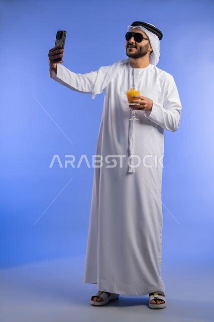 استخدام التقنيات التكنولوجية فى مجال التصوير , الاستمتاع بتصوير الرحلة, الاستمتاع بشرب العصير الطبيعي البارد, بورترية لرجل عربي خليجي اماراتي يرتدي الزي التقليدي يقوم بالتقاط صورة سيلفي عبر الهاتف المحمول, صورة لكامل الجسم, خلفية زرقاء