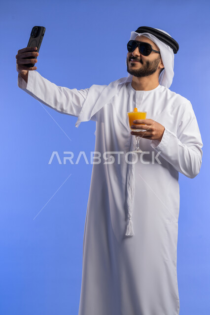اجراء مكالمة فيديو و توثيق الذكريات, الاستمتاع بتصوير الرحلة, الاستمتاع بشرب العصير الطبيعي البارد, بورترية لرجل عربي خليجي اماراتي يرتدي الزي التقليدي يقوم بالتقاط صورة سيلفي عبر الهاتف المحمول, خلفية زرقاء