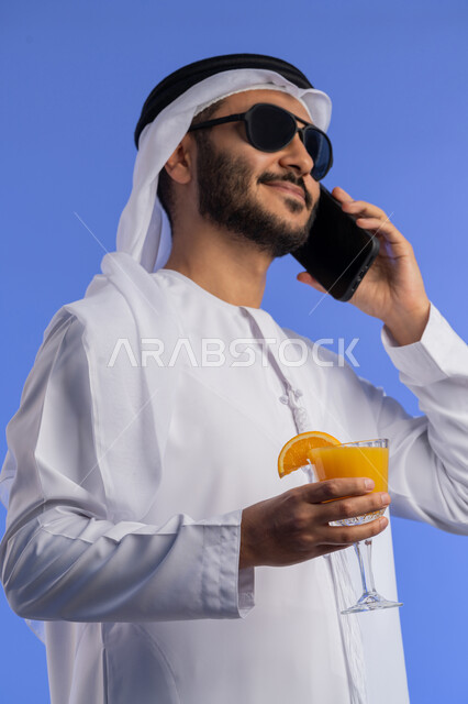 Having a good time in the summer, close-up portrait of a smiling young Arab Emirati man wearing traditional clothes and sunglasses, smiling and talking on the mobile phone with happy gestures, drinking refreshing drinks, blue background