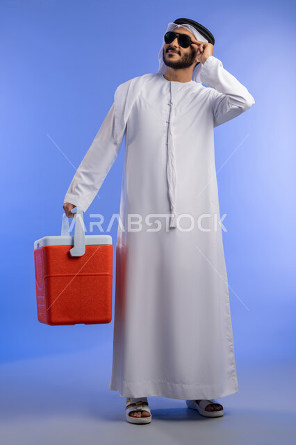 قضاء وقت العطلة الصيفية بالسياحة, بورتريه لشاب عربي خليجي اماراتي مبتسم يرتدي الزي التقليدي والنظارة الشمسية مبتسم يقف و يقوم بالنظر إلى و شيء ما بإيماءات الاسترخاء والراحة و يمسك بيده صندوق تبريد ,صورة لكامل الجسم,خلفية زرقاء