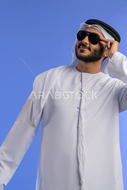 أجواء من الراحة والهدوء تحت شمس الصيف , بورتريه مقرب لشاب عربي خليجي اماراتي مبتسم يرتدي الزي التقليدي والنظارة الشمسية يقف و يقوم بالنظر إلى شيء ما بإيماءات الاسترخاء والراحة, الاستمتاع بالإجازة والمناظر الطبيعيه, خلفية زرقاء
