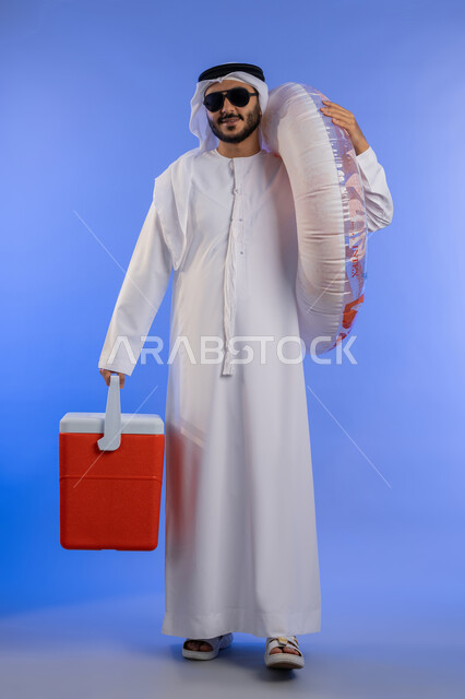 قضاء وقت ممتع في فصل الصيف, بورتريه لشاب عربي خليجي اماراتي مبتسم يرتدي الزي التقليدي والنظارة الشمسية و العوامة استعداد للسباحة في الشاطئ, حافظة البرودة للعصائر, الاستمتاع بالعطلة الصيفية, الجسم بالطول الكامل, خلفية زرقاء