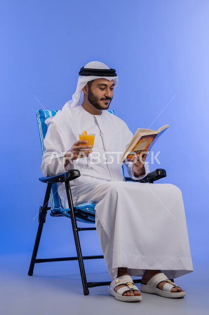 الاستمتاع بالمشروبات الباردة, ممارسة هواية مفيدة في أوقات الفراغ, بورتريه لشاب عربي خليجي اماراتي مبتسم يرتدي الزي التقليدي يجلس على كرسي مريح بتعابير السرور و يقوم بقراءة كتاب ما, قضاء وقت العطلة الصيفية بالسياحة , خلفية زرقاء.
