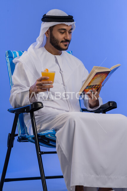 تناول المشروبات الصيفية المنعشة, ممارسة هواية مفيدة فى أوقات الفراغ, بورتريه لشاب عربي خليجي اماراتي مبتسم يرتدي الزي التقليدي  يجلس على كرسي مريح بتعابير السرور و يقوم بقراءة كتاب ما, رحلة شابية ممتعه, قضاء وقت العطلة الصيفية بالسياحة, خلفية زرقاء