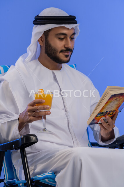 ممارسة الهواية المفضلة فى أوقات الراحة, بورتريه لشاب عربي خليجي اماراتي مبتسم يرتدي الزي التقليدي يقوم بقراءة كتاب ما و هو يجلس على كرسي مريح,الاستمتاع بالمشروبات الباردة،  رحلة شابية ممتعه, قضاء وقت العطلة الصيفية بالسياحة,  خلفية زرقاء