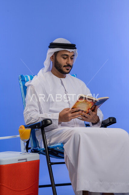 تناول الفواكه الصيفية المنعشة, بورتريه لشاب عربي خليجي اماراتي مبتسم يرتدي الزي التقليدي يقوم بقراءة كتاب ما و هو يجلس على كرسي بجانب صندوق تبريد, رحلة شابية ممتعه, قضاء وقت العطلة الصيفية بالسياحة, خلفية زرقاء
