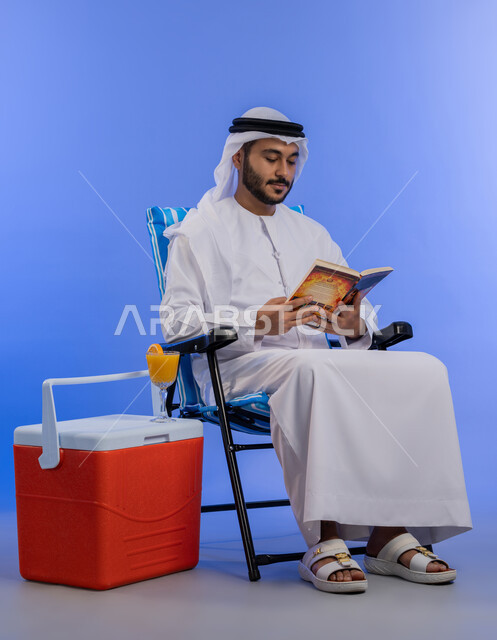 الاستمتاع بالمشروبات الباردة, بورتريه لشاب عربي خليجي اماراتي مبتسم يرتدي الزي التقليدي يقوم بقراءة كتاب ما و هو يجلس على كرسي بجانب صندوق تبريد, رحلة شابية ممتعه, قضاء وقت العطلة الصيفية بالسياحة, خلفية لكامل الجسم , خلفية زرقاء