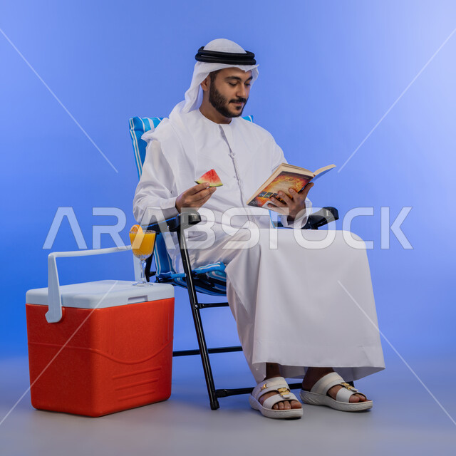 قضاء وقت العطلة الصيفية بالسياحة, تناول الفواكه الصيفية المنعشة, بورتريه لشاب عربي خليجي اماراتي مبتسم يرتدي الزي التقليدي يقوم بقراءة كتاب ما و هو يجلس على كرسي بجانب صندوق تبريد, رحلة شابية ممتعه, خلفية لكامل الجسم, خلفية زرقاء