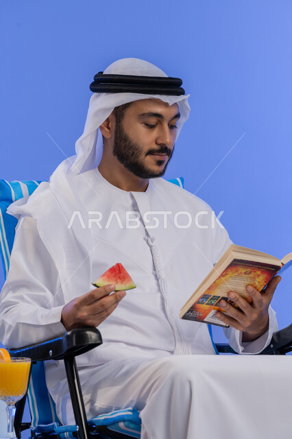 ممارسة هواية مفيدة فى أوقات الفراغ ,تناول الفواكه الصيفية المنعشة, بورتريه لشاب عربي خليجي اماراتي مبتسم يرتدي الزي التقليدي يقوم بقراءة كتاب ما و هو يجلس على كرسي مريح , رحلة شابية ممتعه, قضاء وقت العطلة الصيفية بالسياحة , خلفية زرقاء