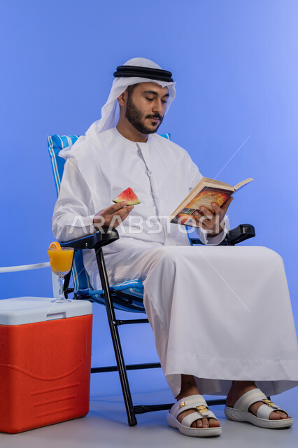 تناول الفواكه الصيفية المنعشة, بورتريه لشاب عربي خليجي اماراتي مبتسم يرتدي الزي التقليدي يقوم بقراءة كتاب ما و هو يجلس على كرسي بجانب صندوق تبريد, رحلة شابية ممتعه, قضاء وقت العطلة الصيفية بالسياحة, خلفية لكامل الجسم , خلفية زرقاء