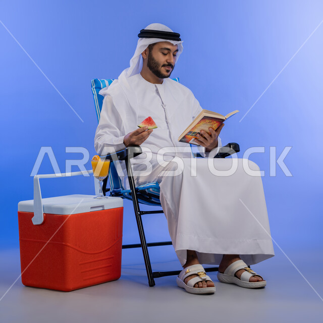 ممارسة هواية مفيدة في أوقات الفراغ, بورتريه لشاب عربي خليجي اماراتي مبتسم يرتدي الزي التقليدي يقوم بقراءة كتاب ما و هو يجلس على كرسي بجانب صندوق تبريد, رحلة شابية ممتعه, تناول الفاكهه الصيفية المنعشه, خلفية لكامل الجسم , خلفية زرقاء