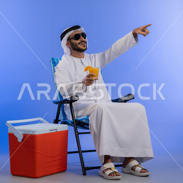 رحلة شبابية ممتعه, تناول المشروبات المنعشة, بورتريه لشاب عربي خليجي اماراتي مبتسم يرتدي الزي التقليدي يقوم بالإشارة الى شيء ما و هو يجلس على كرسي بجانب صندوق تبريد, قضاء وقت العطلة الصيفية بالسياحة, خلفية لكامل الجسم, خلفية زرقاء