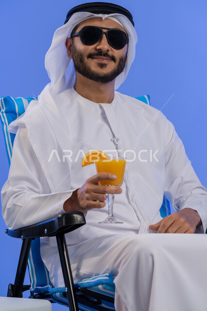 فعاليات وأنشطة ترفيهية سياحية, الاستمتاع بالمشروبات الباردة فى الأجواء الحارة, بورتريه لشاب عربي خليجي اماراتي مبتسم يرتدي الزي التقليدي والنظارة الشمسية ينظر الى الكاميرا يقوم بشرب عصير البرتقال الطبيعى و هو يجلس على كرسي مريح, خلفية زرقاء