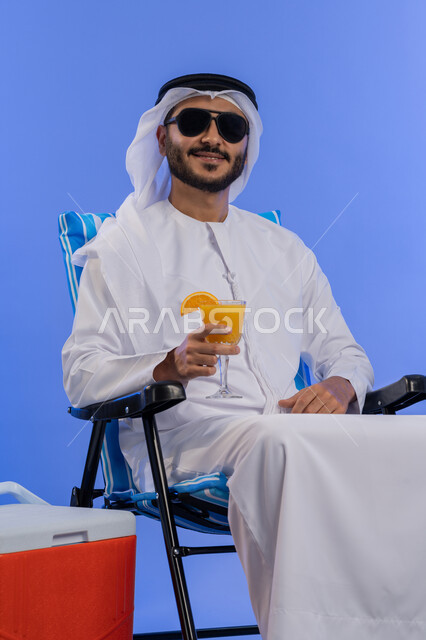 الخروج برحلة سياحية فى أوقات الفراغ, قضاء اجازة الصيف بالاماكن السياحية, بورتريه لشاب عربي خليجي اماراتي مبتسم يرتدي الزي التقليدي والنظارة الشمسية ينظر الي الكاميرا و يقوم بشرب عصير البرتقال الطبيعى و هو يجلس على كرسي مريح بجانبة حافظة تبريد , خلفية زرقاء