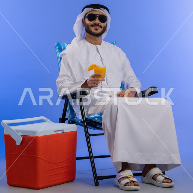 تحسين المزاج وإعطاء شعور بالانتعاش, بورتريه لشاب عربي خليجي اماراتي مبتسم يرتدي الزي التقليدي والنظارة الشمسية يقوم بشرب عصير البرتقال الطبيعى و هو يجلس على كرسي مريح بجانبة حافظة تبريد, صورة لكامل الجسم , خلفية زرقاء