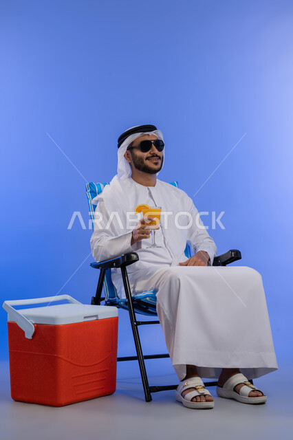 الخروج برحلة سياحية في أوقات الفراغ , بورتريه لشاب عربي خليجي اماراتي مبتسم يرتدي الزي التقليدي والنظارة الشمسية يقوم بشرب عصير البرتقال الطبيعى وهو يجلس على كرسي مريح بجانبة حافظة تبريد,رحلة شبابية ممتعه, صورة  لكامل الجسم، خلفية زرقاء