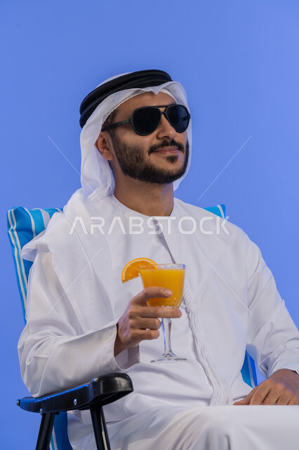 قضاء أيام ممتعة فى العطلة , الخروج برحلة سياحية فى أوقات الفراغ, بورتريه مقرب لشاب عربي خليجي اماراتي مبتسم يرتدي الزي التقليدي والنظارة الشمسية يقوم بشرب عصير البرتقال الطبيعى و هو يجلس على كرسي مريح, رحلة شبابية ممتعه , خلفية زرقاء