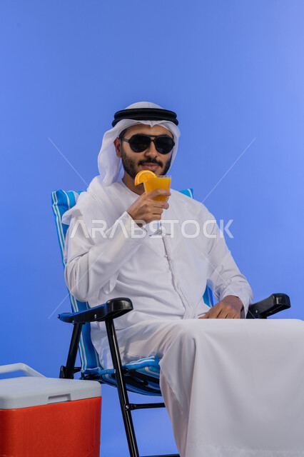 الاستمتاع بالمشروبات الصيفية الباردة , الخروج برحلة سياحية فى أوقات الفراغ, بورتريه لشاب عربي خليجي اماراتي مبتسم يرتدي الزي التقليدي والنظارة الشمسية يقوم بشرب عصير البرتقال الطبيعى و هو يجلس على كرسي مريح بجانب حافظة تبريد, خلفية زرقاء