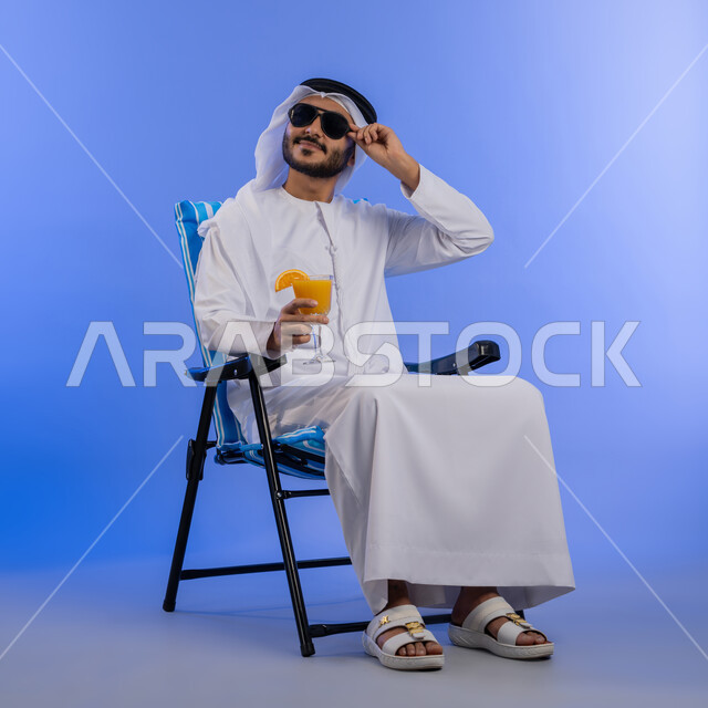 Fun youth trip, enjoying cold drinks, going on a tourist trip in free time, portrait of a smiling Emirati Gulf Arab young man wearing traditional clothing and sunglasses sitting on a comfortable chair looking at something, full body photo, blue background