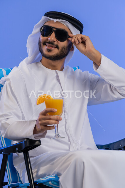 الاستمتاع بالمشروبات الباردة, الخروج برحلة سياحية فى أوقات الفراغ, بورتريه مقرب لشاب عربي خليجي اماراتي مبتسم يرتدي الزي التقليدي والنظارة الشمسية يجلس على كرسي مريح ينظر الى شيء ما,رحلة شبابية ممتعه, خلفية زرقاء