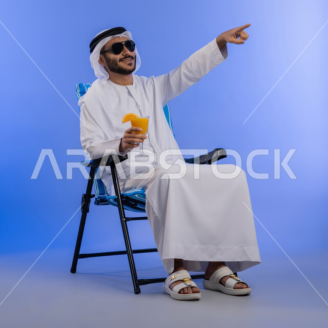 الخروج برحلة سياحية فى أوقات الفراغ, بورتريه لشاب عربي خليجي اماراتي مبتسم يرتدي الزي التقليدي والنظارة الشمسية  يجلس على كرسي مريح و يشير الى شيء ما,  الاستمتاع بالمشروبات الباردة, صورة للجسم الكامل, خلفية زرقاء