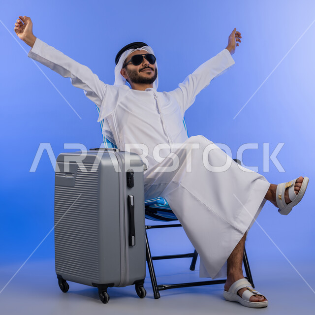 قضاء العطلة الصيفية فى المنتجعات السياحية , بورتريه لشاب عربي خليجي اماراتي مبتسم يرتدي الزي التقليدي والنظارة الشمسية يجلس على كرسي مريح, رجل اماراتي يقوم برفع ذراعية بإيماءات الاسترخاء والراحة ,صورة لكامل الجسم, خلفية زرقاء