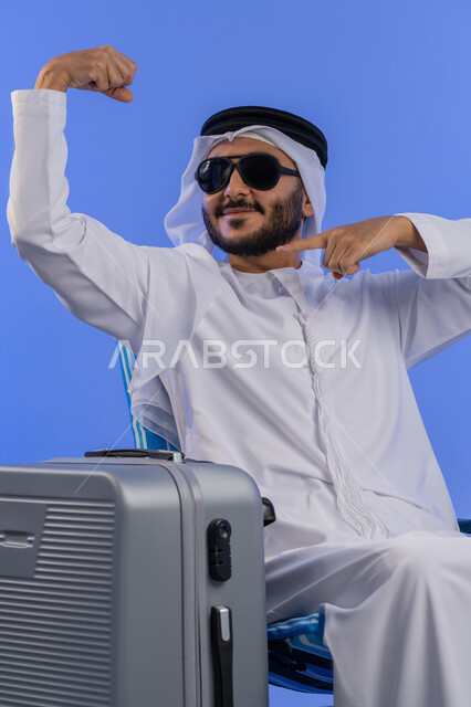 الذهاب فى رحلة سياحية ممتعة، فعاليات وأنشطة ترفيهية سياحية, مفهوم السياحة فى إجازة الصيف، بورترية  لشاب عربي خليجي  اماراتي يرتدي الزى التقليدي يجلس بجانب شنطه سفر و يقوم برفع ذراعية بايماءات وجه ويدين بتعابير الثقة والاستعداد, خلفية زرقاء.