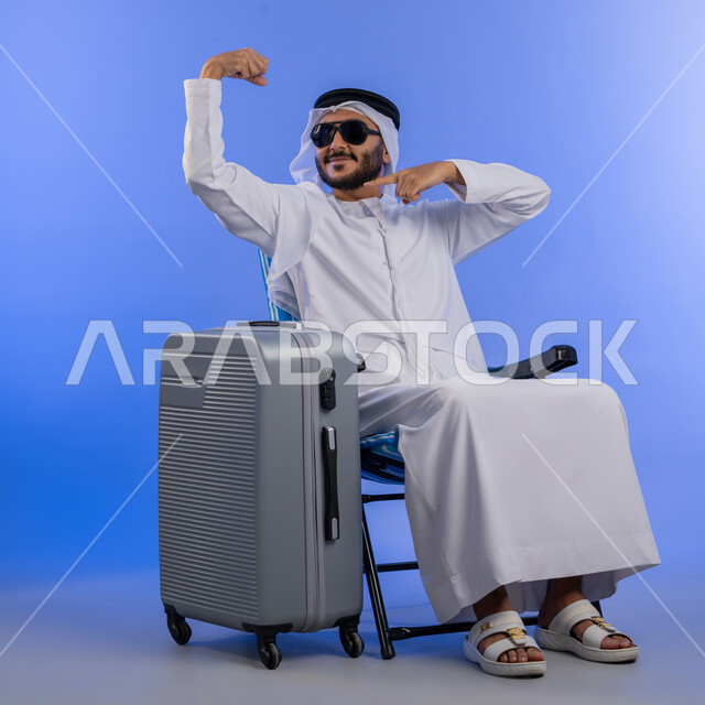 Preparing for summer vacation, portrait of a young Emirati Gulf Arab man wearing traditional dress sitting next to his suitcase and raising his arms with facial and hand gestures expressing confidence and readiness, full body photo, blue background.