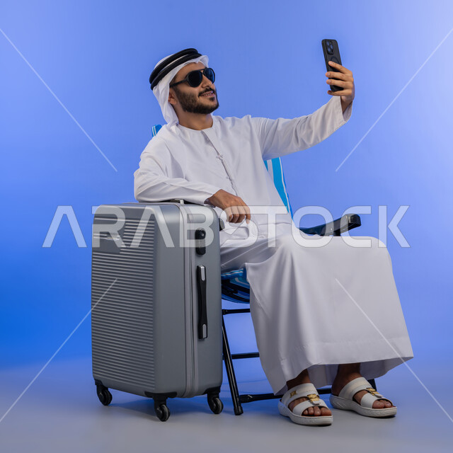 الاستعداد للسفر بإيماءات السعادة , بورترية لرجل عربى خليجي امراتي يرتدي الغتره البيضاء و الكندورة و النظارة الشمسية يجلس على كرسي بجانبة شنطه سفر يقوم بالتقاط صورة سيلفي عبر الهاتف المحمول , صورة لكامل الجسم , خلفية زرقاء