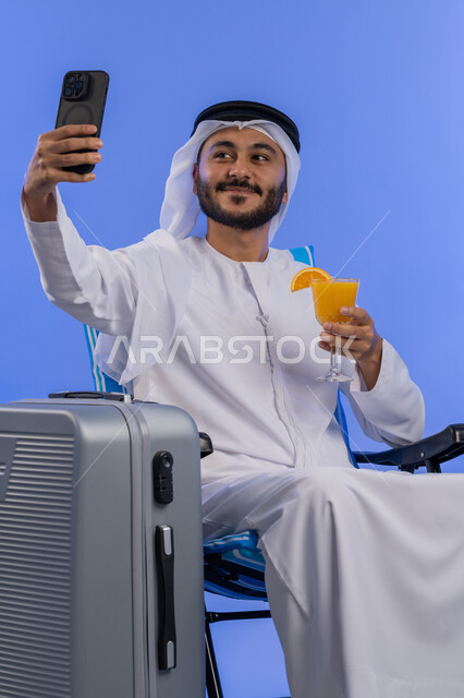 التقاط الصور التذكارية وتوثيق اللحظات السعيدة , بورترية لرجل عربى خليجي امراتي يرتدي الغتره البيضاء و الكندورة يجلس علي كرسي بجانبة شنطه سفر يقوم بالتقاط صورة سيلفي عبر الهاتف المحمول , شرب العصائر المنعشة , خلفية زرقاء.